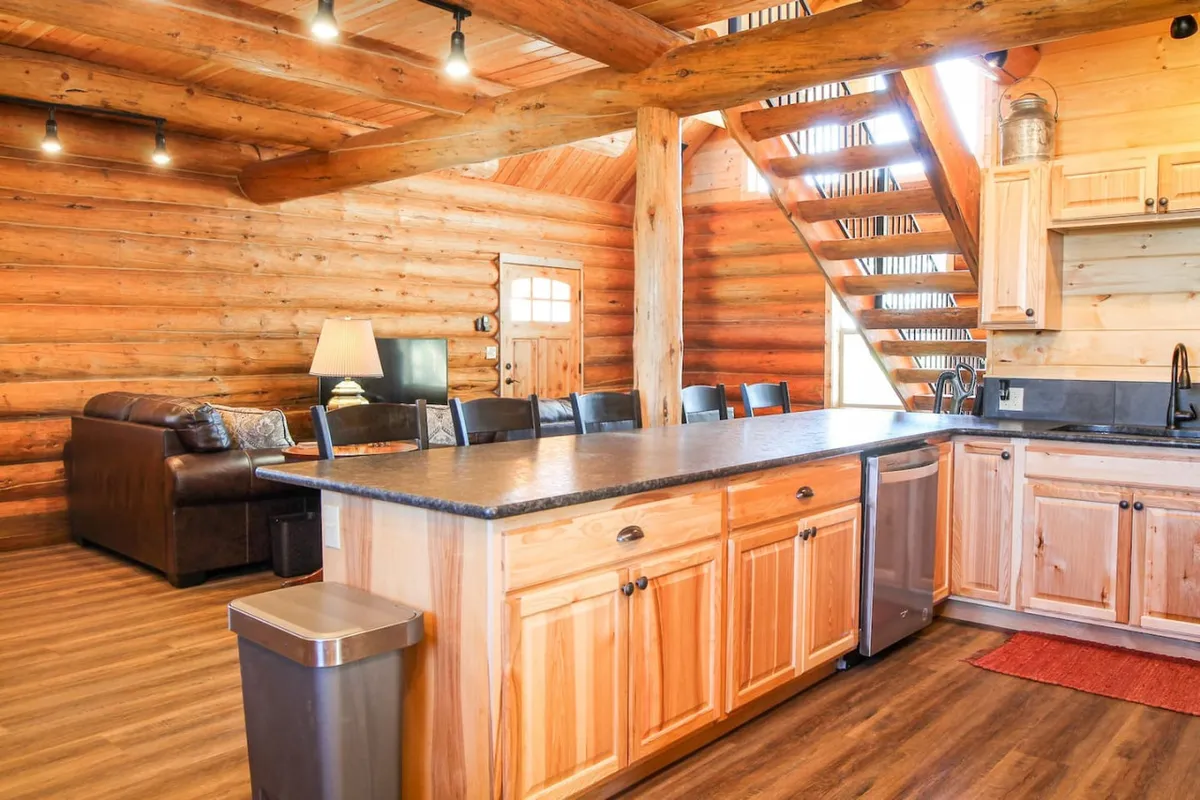 This interior view captures an open-plan living space with visible wooden beams, a central kitchen island, a sofa, and a staircase. The layout encourages connection among the kitchen, dining, and living areas, with warm lighting creating a welcoming ambiance ideal for gatherings and shared moments.
