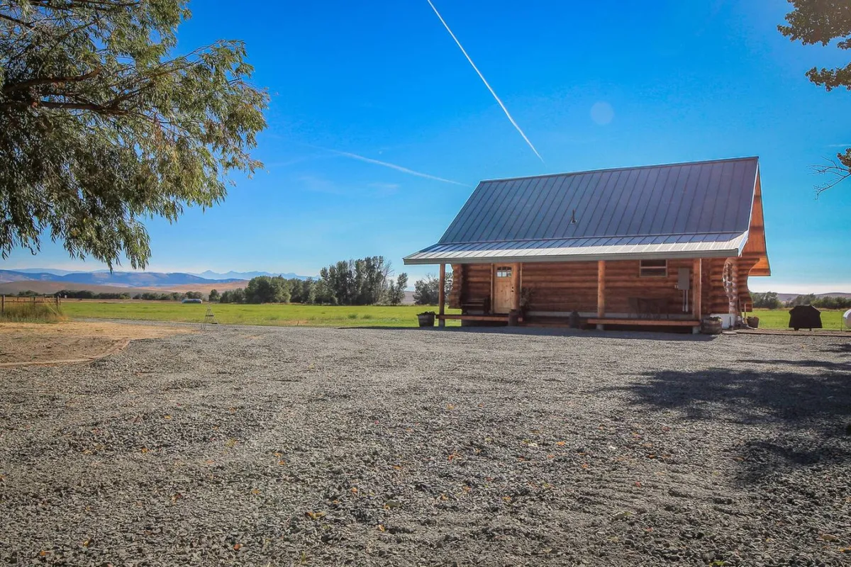 A wide-angle shot of the log cabin set against open fields with the mountain range in the distance. The foreground reveals the deck area that blends into the surrounding ranch landscape, inviting viewers to imagine the expansive views and tranquil setting the decks provide.