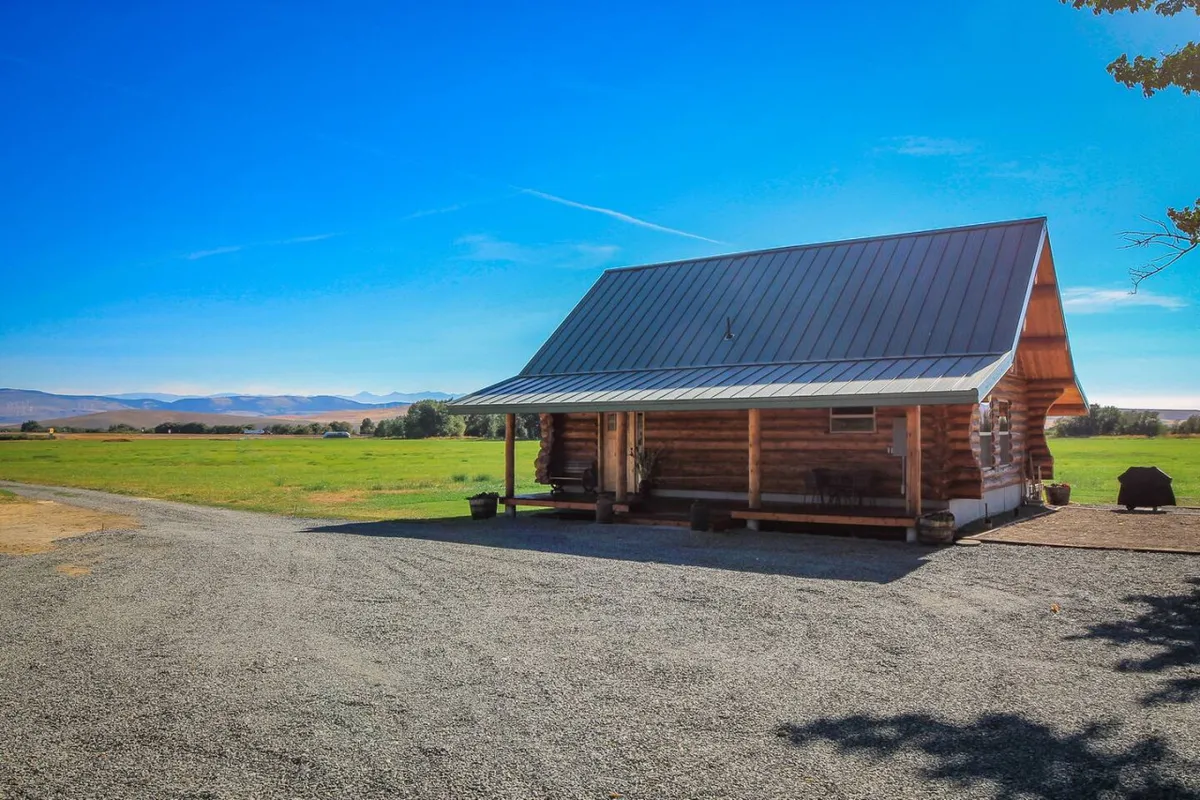 This exterior image shows the Hill Cabin nestled amid a peaceful landscape, with a gravel driveway and expansive surroundings. The log cabin is featured prominently, blending into the natural setting. The open land hints at a rural, adventure-ready environment, perfect for launching into nearby activities.