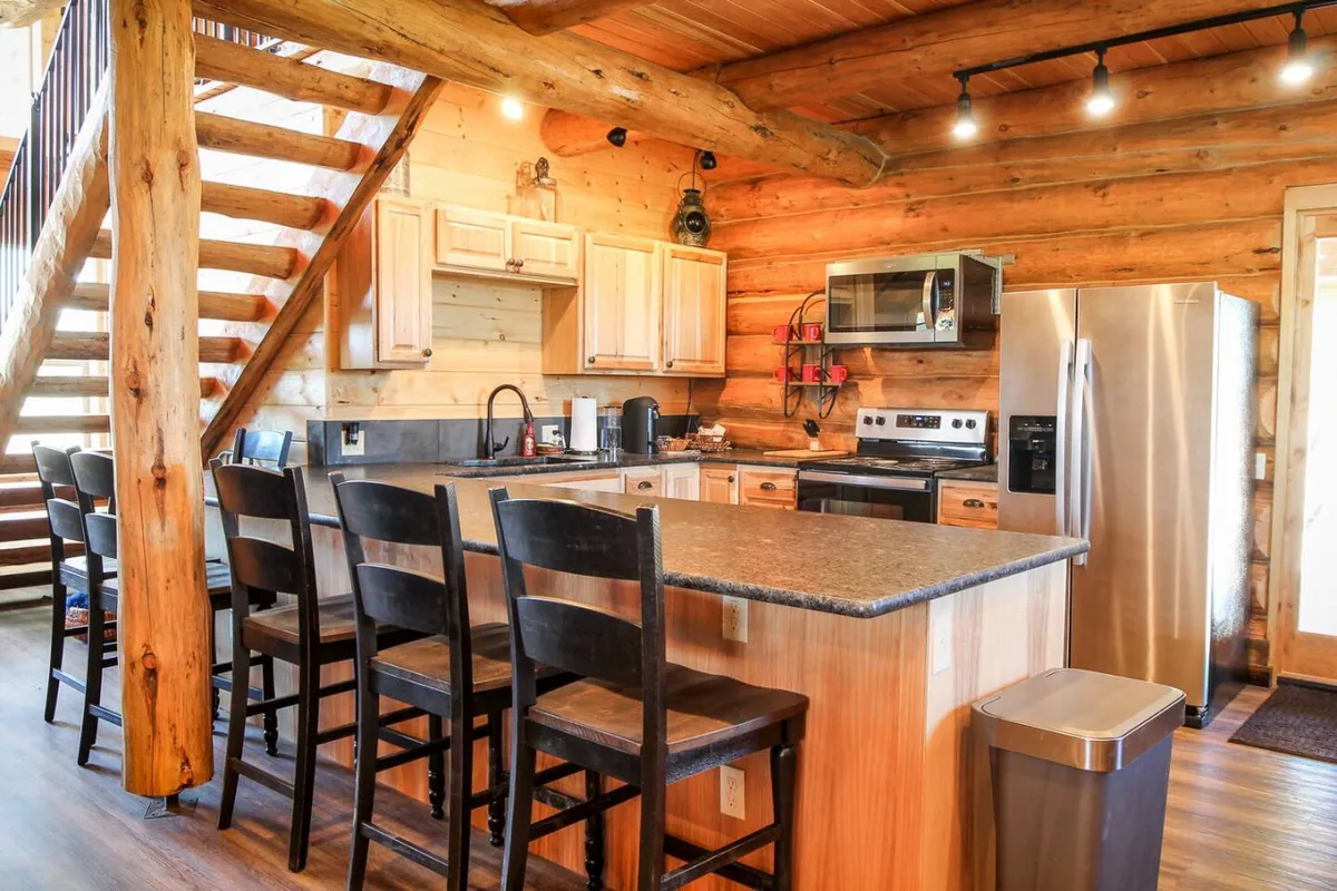 This image presents a warm, rustic kitchen with a central island, stainless steel appliances, wooden cabinetry, and log walls. Sunlight filters through, illuminating a space perfect for cooking, gathering, and casual dining. The visible appliances and inviting layout highlight the gourmet possibilities of the kitchen.