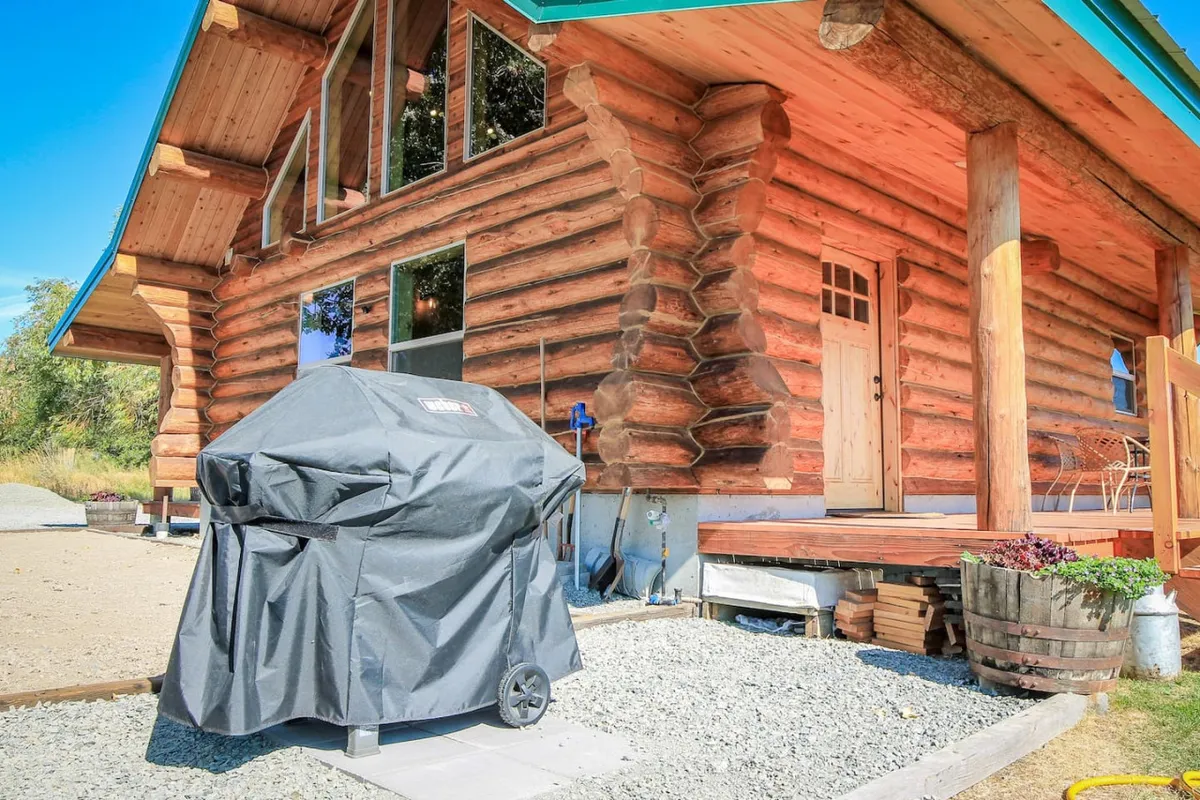 A log cabin exterior with a welcoming front porch, grill, and several windows. The porch offers a spot for outdoor dining or savoring a fresh morning coffee, while the grill hints at the potential for shared meals. The scene is inviting, with sunlight highlighting the warm wooden textures.