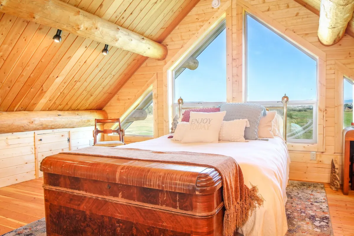 The image shows a cozy, inviting bedroom interior with a plush bed adorned with decorative pillows, warm wooden beams overhead, and expansive windows framing stunning views of the Elkhorn Mountains and rolling pastures. Natural light floods the space, creating a tranquil atmosphere perfect for unwinding.