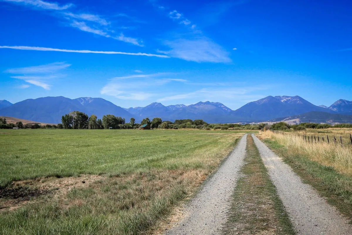 A sweeping view of a gravel road leading toward a majestic mountain range under an open blue sky. Green fields border the road, creating a sense of openness and natural beauty. The scene invites exploration and promises breathtaking sunrises or sunsets over the Elkhorn Mountains.