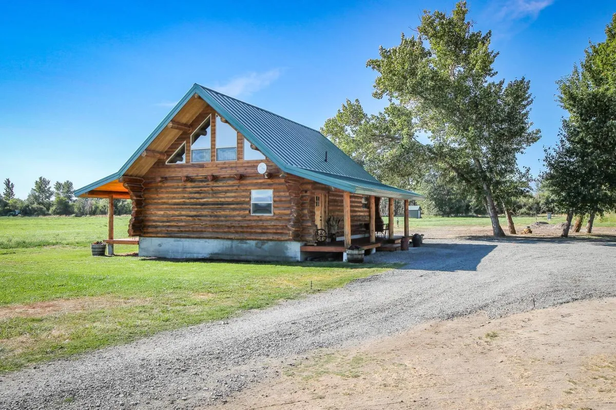 The log cabin with a green roof is surrounded by lush greenery and a gravel driveway. The porch seating invites relaxation while the landscape beckons for nature walks and outdoor gatherings. The scene feels tranquil and welcoming, emphasizing the property’s connection to its surroundings.