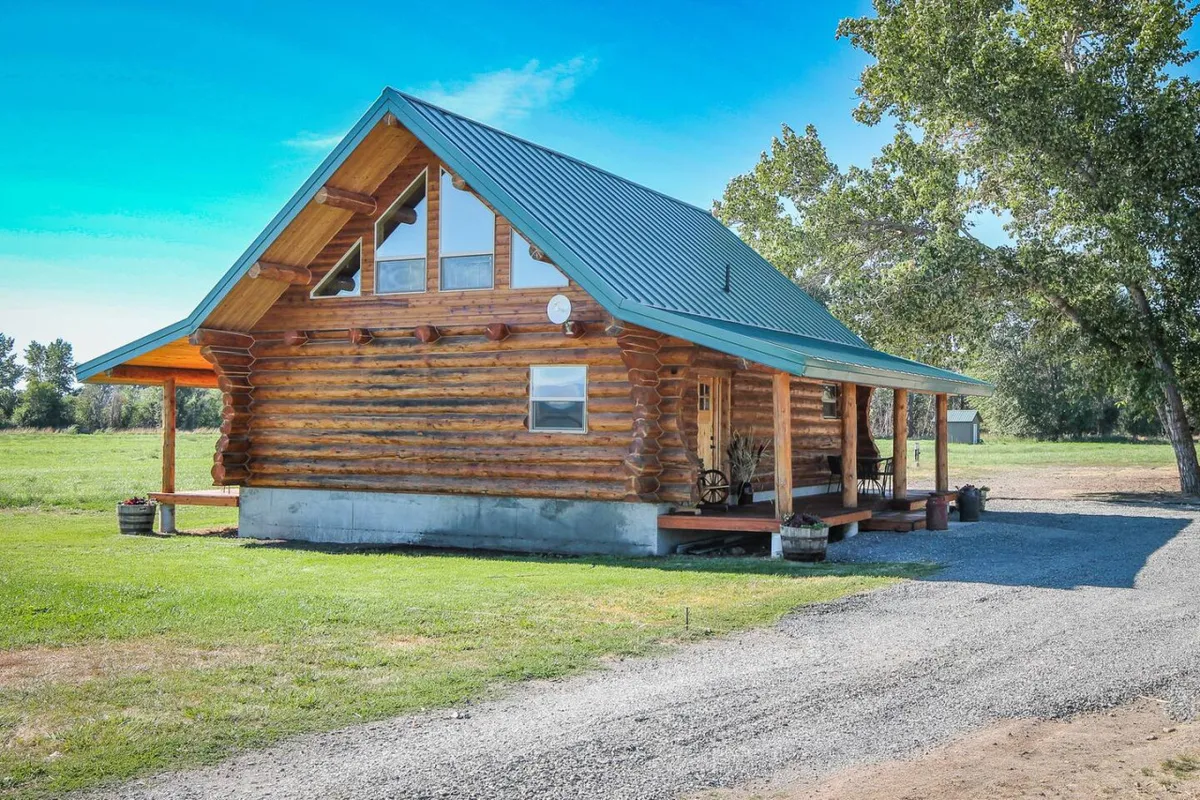 A cozy log cabin with a green metal roof sits nestled in a landscaped yard. The front porch is adorned with wooden rocking chairs, inviting relaxation and quiet moments. The scene exudes tranquility, with lush greenery surrounding the cabin and ample space for enjoying the outdoors.