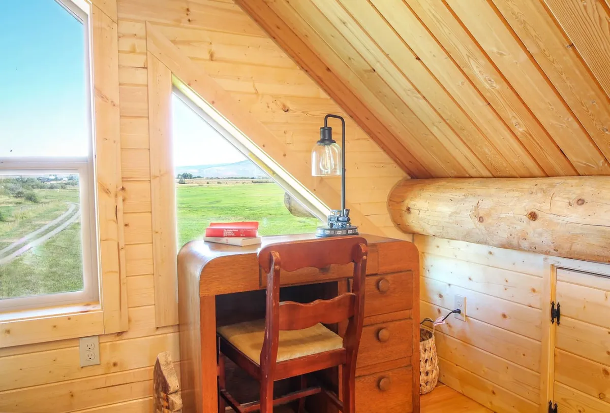 A tranquil nook with a wooden desk, a lamp casting a gentle glow, and a window offering a quiet view of the natural landscape. Books are neatly arranged, inviting quiet moments of reflection and relaxation.