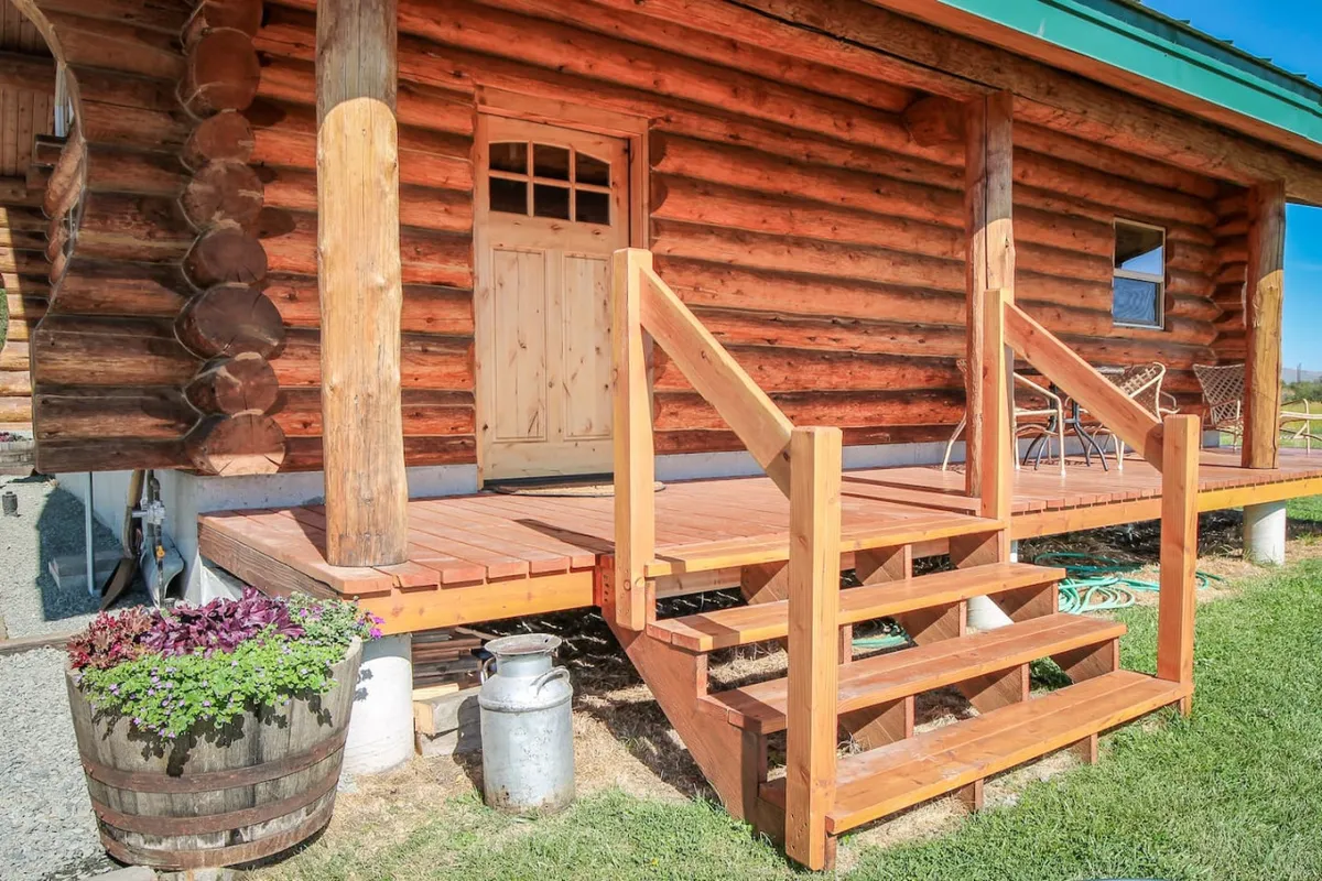 A beautifully renovated log cabin with an inviting wooden stairway, a welcoming doorway, and a cozy outdoor seating area adorned with a flower pot. The porch offers ample space for relaxing and soaking in the surrounding nature.