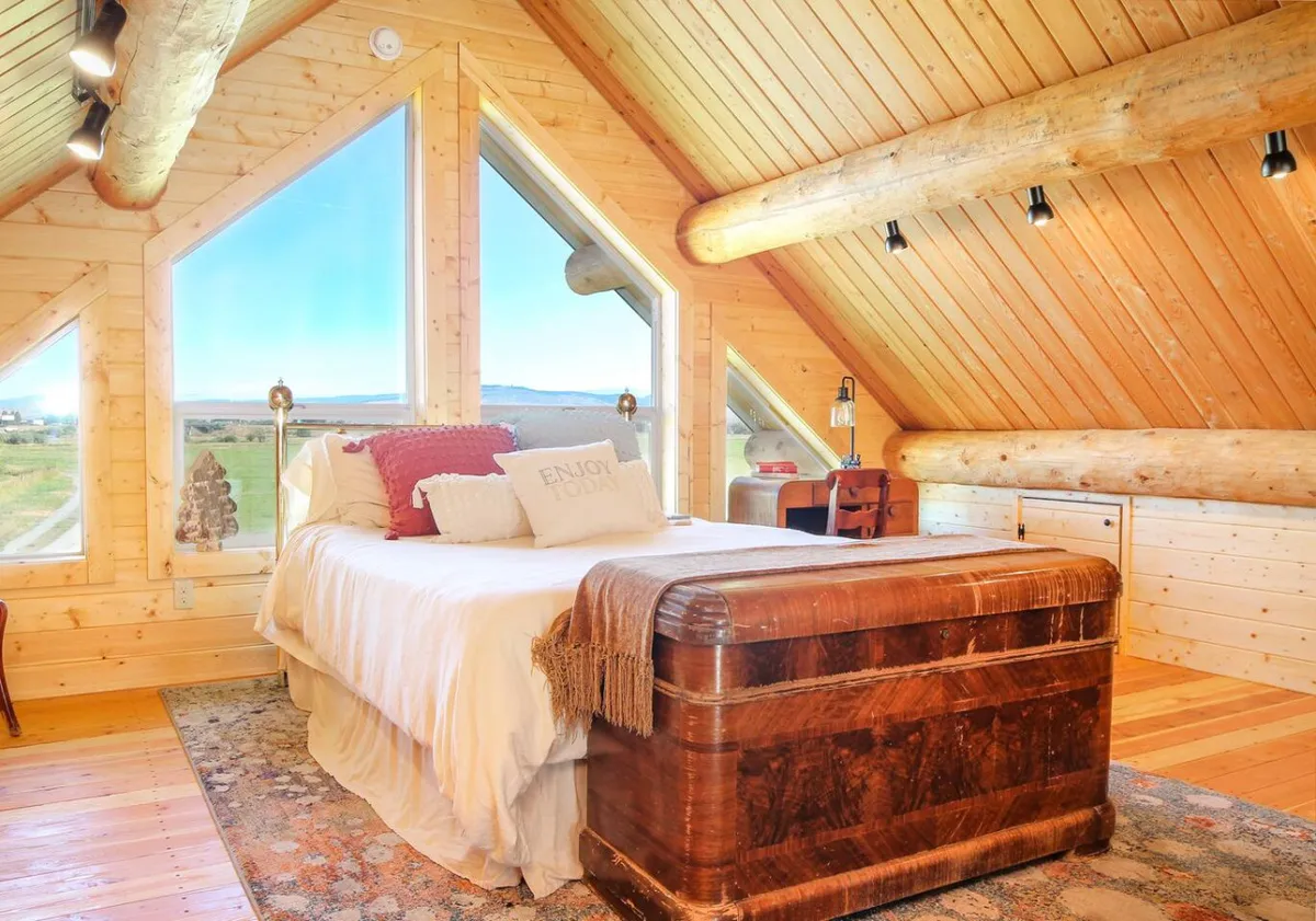 A serene upstairs bedroom featuring a cozy bed with decorative pillows, expansive windows that let in abundant natural light, rustic wooden beams overhead, and an antique trunk at the foot of the bed. Outside the windows, glimpses of the Elkhorn Mountains and rolling pastures enhance the tranquil atmosphere.