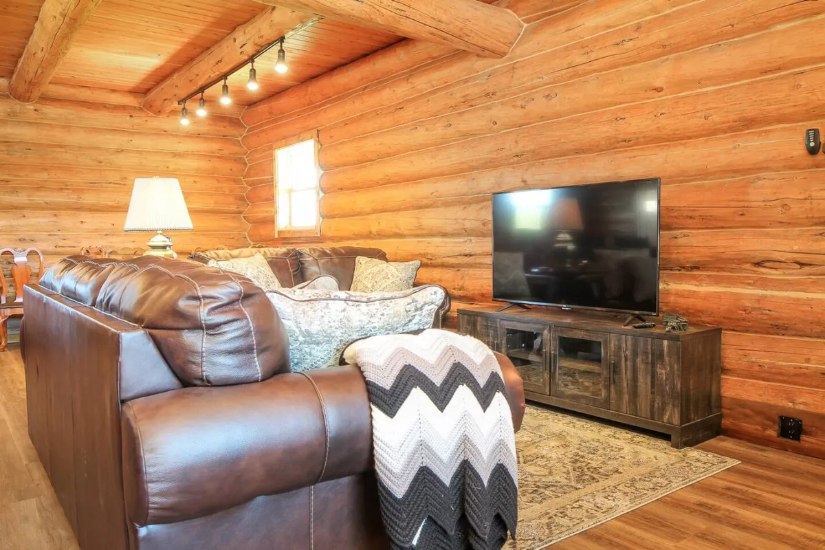 A cozy living area is depicted with a leather sofa draped in a textured throw blanket, a television, and classic wooden walls. The arrangement offers a comfortable spot for watching TV or relaxing after outdoor activities, embodying the inviting and tranquil feel central to the section.
