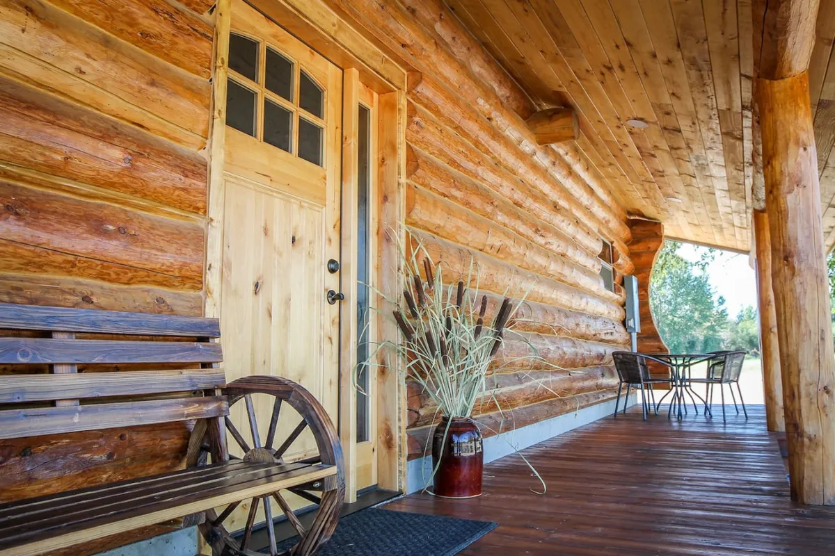 A charming wooden porch features a wheeled bench, potted plant, and comfortable outdoor seating—ideal for enjoying morning coffee or relaxing at sunset. The view hints at the open spaces and rolling pastures beyond.